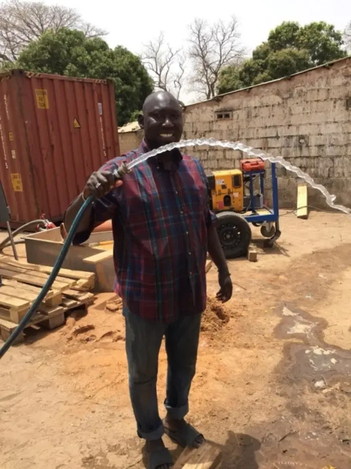 Mann in Gambia hält lachend einen Schlauch, aus dem das Wasser des neuen Brunnens sprudelt
